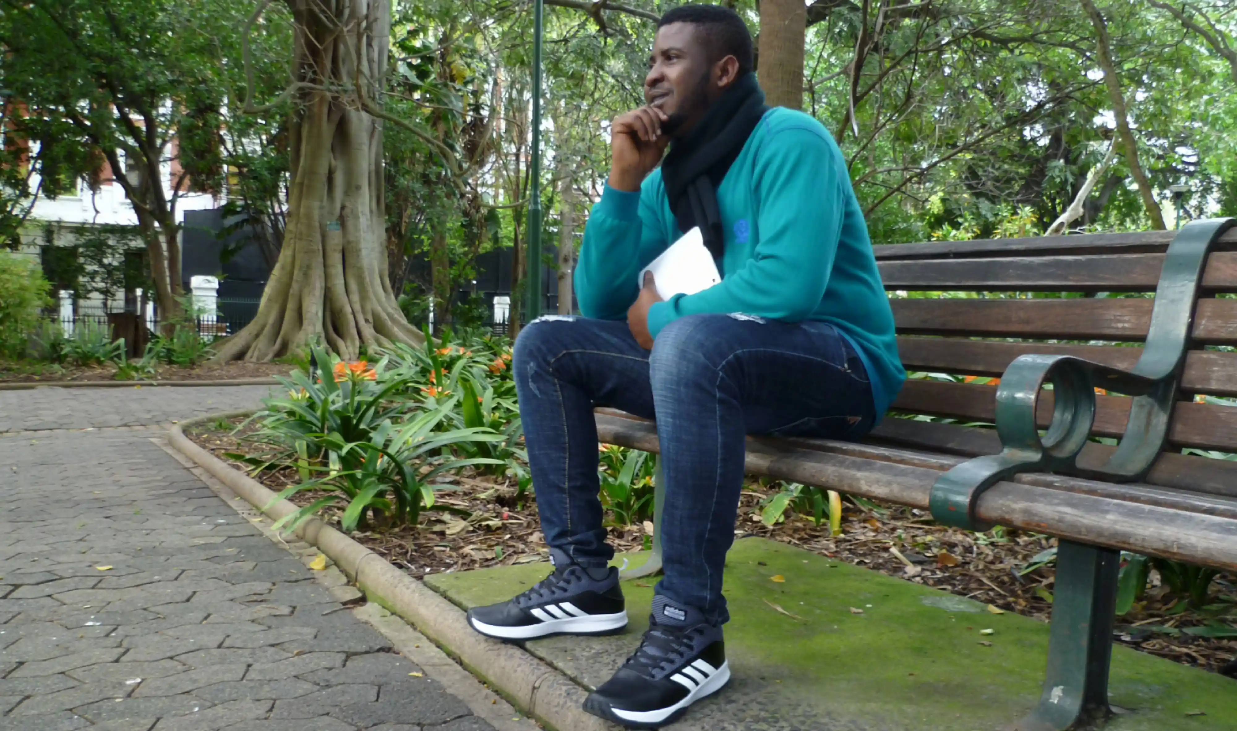 Nzube Okafor sitting on an outdoor bench in Company's Garden, Cape Town, wearing a teal sweatshirt, dark jeans, a black scarf, and Adidas sneakers.